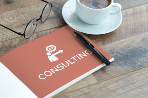 a stock photo of a wooden table. On top of the table are black round glasses, coffee in a white cup on a white saucer, and a red booklet that says consulting with a pen on top. 