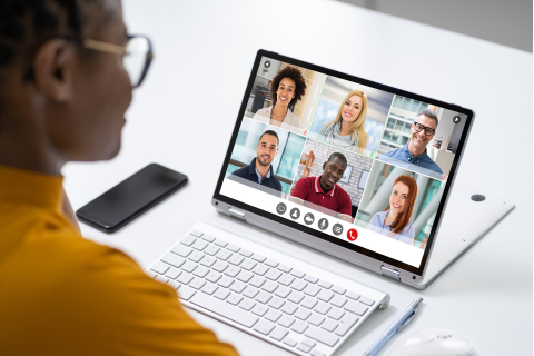 a stock photo of a woman starting at her tablet device on a white table. The tablet shows  six different people on a videoconference call on the screen. Next to the tablet is a keyboard, black phone, pen, and mouse. The lady is wearing a yellow shirt and round glasses.