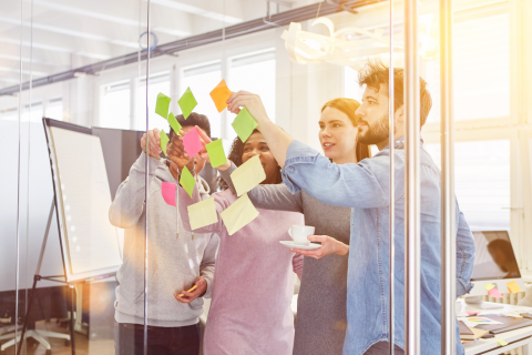 a stock photo of four people brainstorming in a meeting room with glass walls. On the glass wall in front of the group they have places different colored post it notes. In the background there is a desktop, flipchart, and window shining sunlight. One of the group members is holding a white coffee cup and saucer.